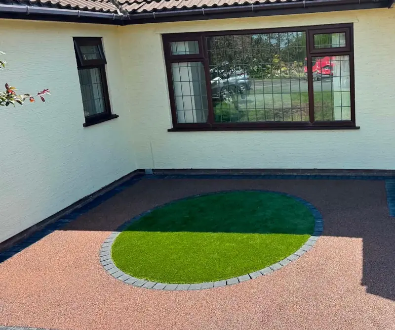 Red resin driveway with a circular artificial lawn edged in charcoal blocks in front of a cream rendered bungalow
