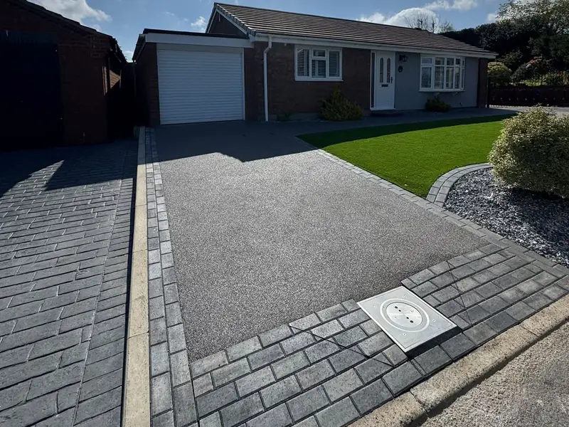 Dark grey resin driveway with a charcoal block paved border, a recessed manhole cover and an artificial lawn in front of a bungalow
