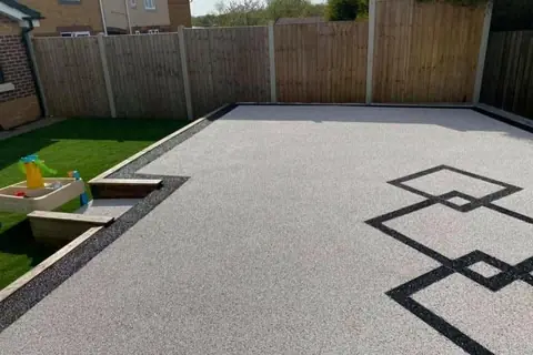 Silver resin surface in a back garden with a black interlocking diamond pattern and sleeper steps down to an artificial lawn