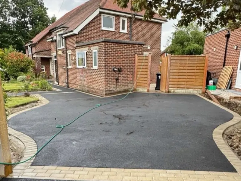 New black tarmac driveway with a curved buff block paved border beside a red brick house